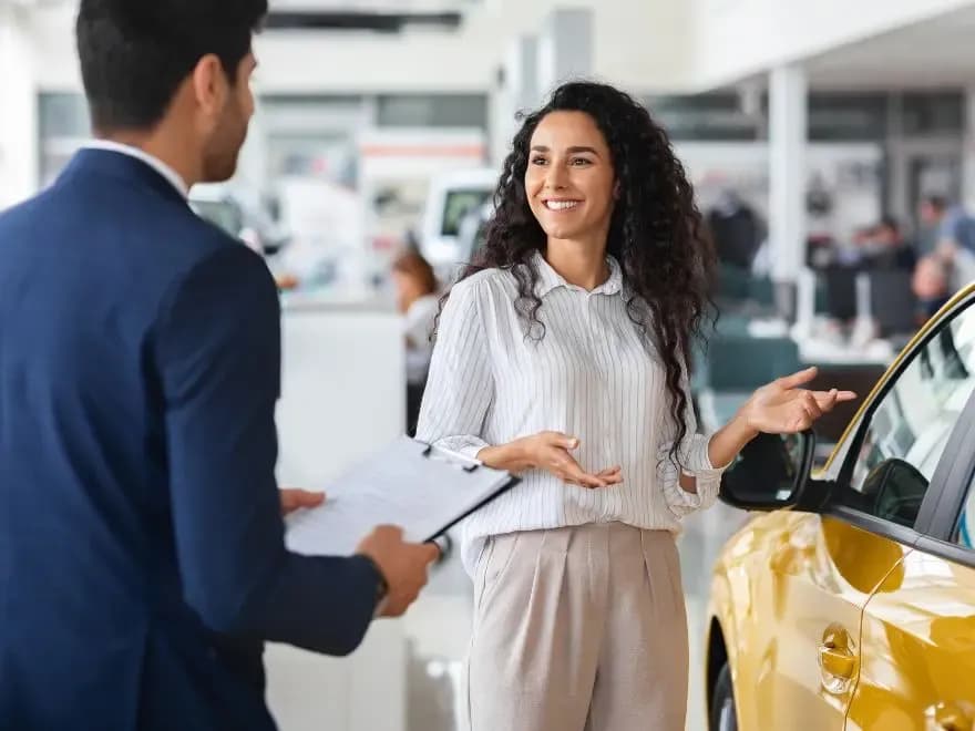 Vendedora de carros gesticulando para um carro amarelo enquanto conversa com um consultor de vendas que segura uma prancheta em uma concessionária, ilustrando o treinamento de equipe para saber como vender carro automático.