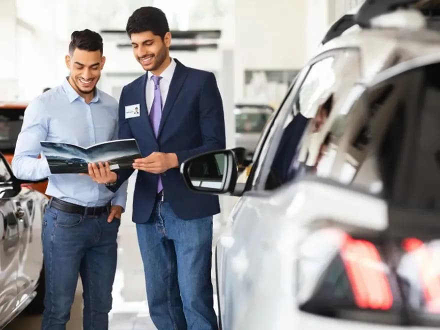 Dois homens em pé ao lado de um carro, sorrindo e olhando para um catálogo de carros aberto, ilustrando uma como saber vender carro automático.