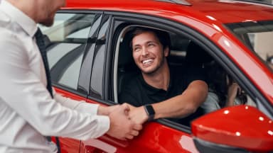 Homem dentro de um carro vermelho apertando a mão de um funcionário da concessionária, formalizando uma venda de um carro com quilometragem alta.